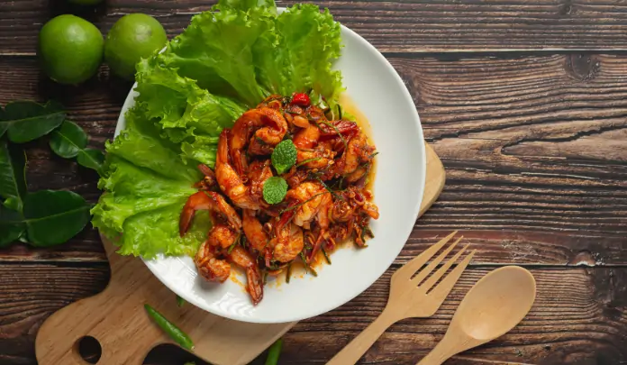 Spicy Indo-Chinese shrimp served on lettuce leaves with herbs on a white plate over a wooden table.