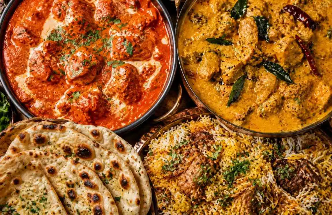 Overhead view of an Indian banquet table with butter chicken, Kerala coconut curry, mutton biryani, and assorted breads shared among a group